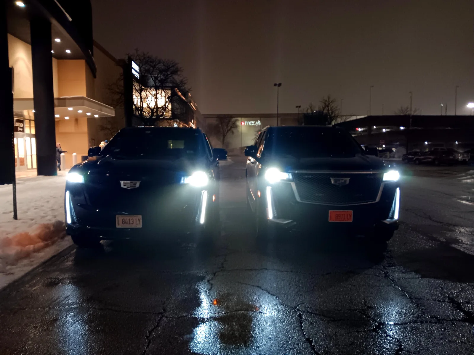Two black Cadillac Escalades with headlights on, parked on wet pavement at night